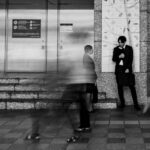 モノクロの駅構内。行き交う人々が長時間露光でぶれて透けるように流れる中、スーツ姿の男性がひとり壁にもたれ、動かずに佇んでいる。