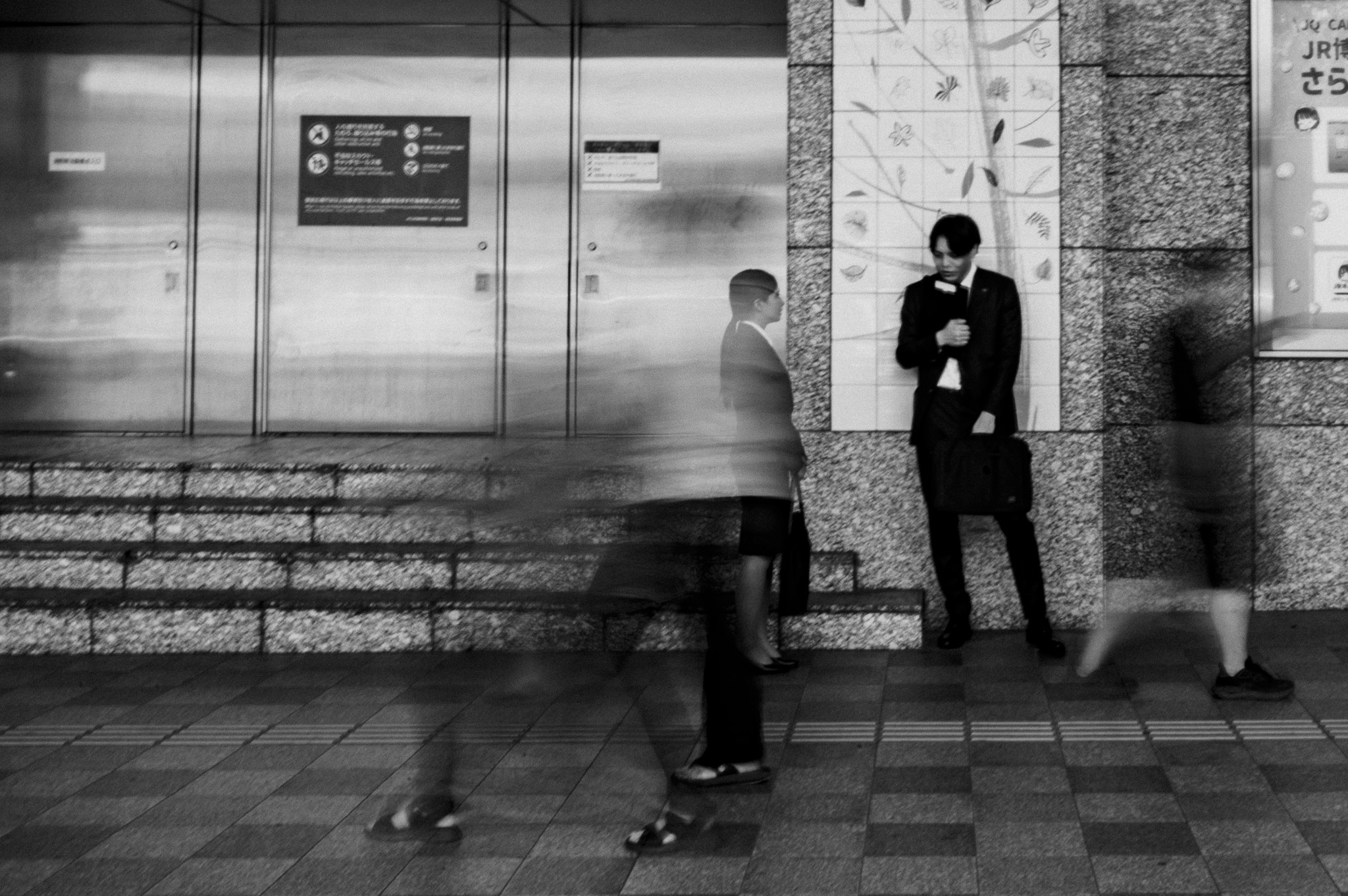 モノクロの駅構内。行き交う人々が長時間露光でぶれて透けるように流れる中、スーツ姿の男性がひとり壁にもたれ、動かずに佇んでいる。