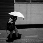 モノクロの街角。強い陽射しの中を、白い日傘を差し、リュックと肩掛けの荷物を持った人が、足元に濃い影を落として歩いている。背景は波板シャッターとコンクリートの壁。路面に「自転車放置禁止区域／熊本市」の標示。